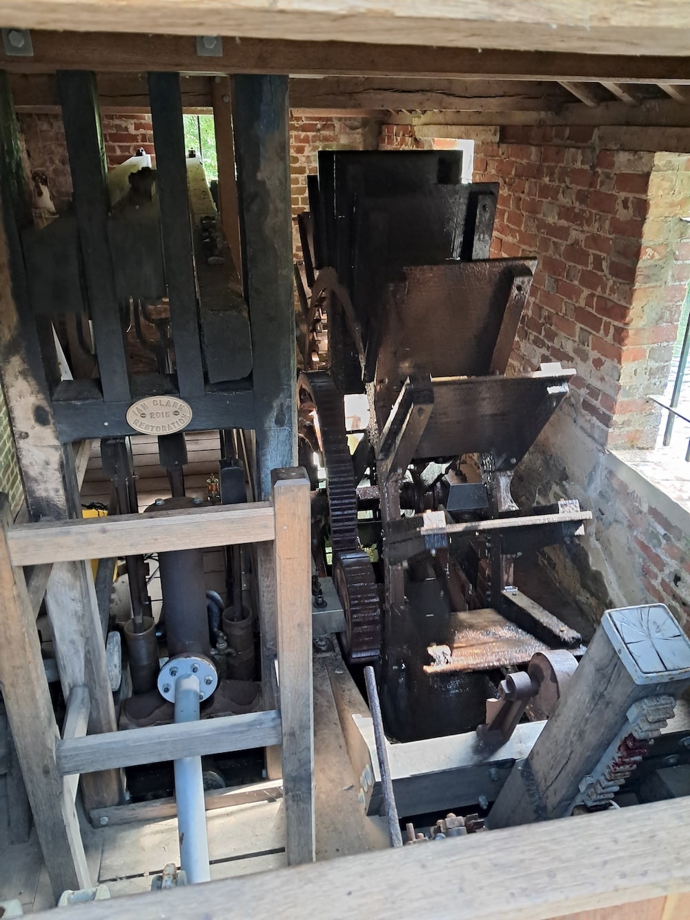 A wooden and metal waterwheel mechanism is seen from above inside a brick-walled historic building.