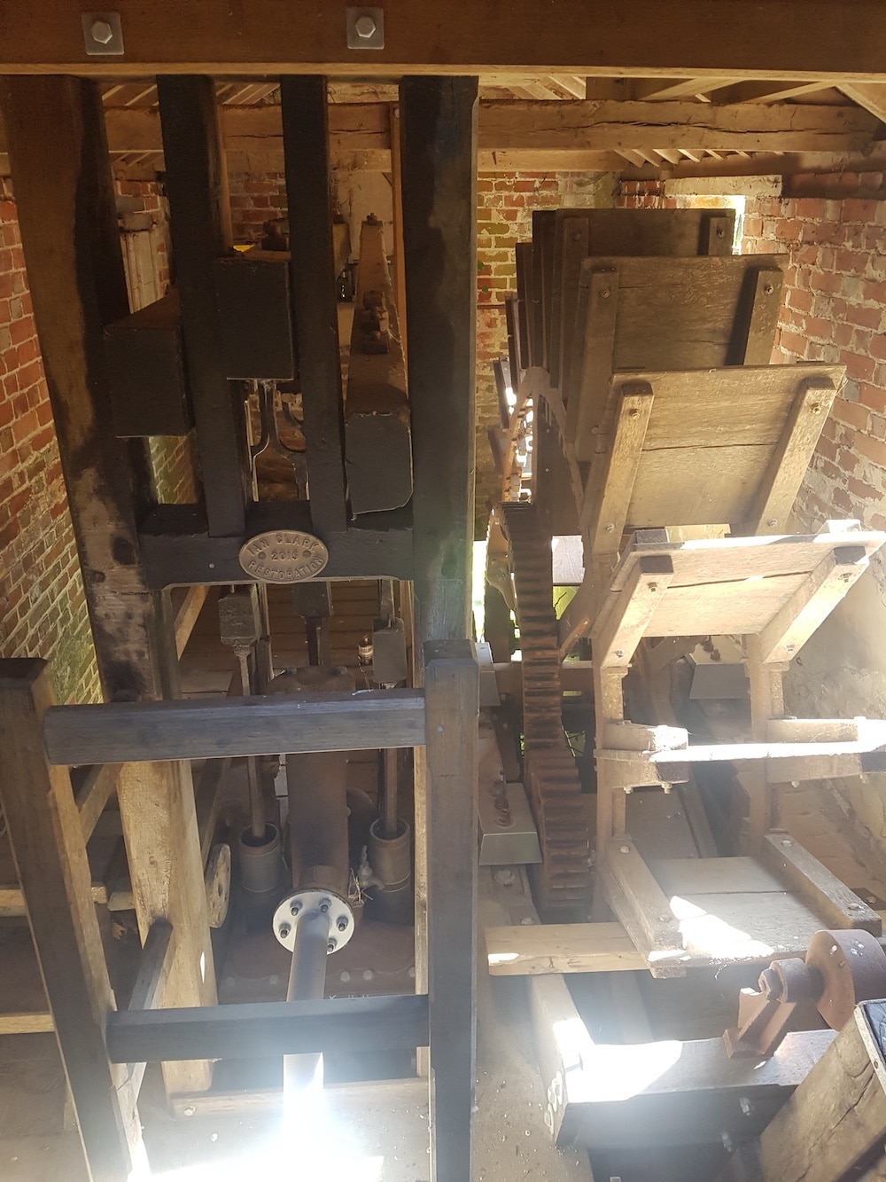 Sunlight streams into a brick building, illuminating wooden gears and beams of an old water-powered mill mechanism.