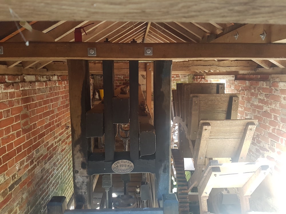 Wooden machinery stands inside a brick building, with beams overhead and sunlight streaming in through an open doorway.