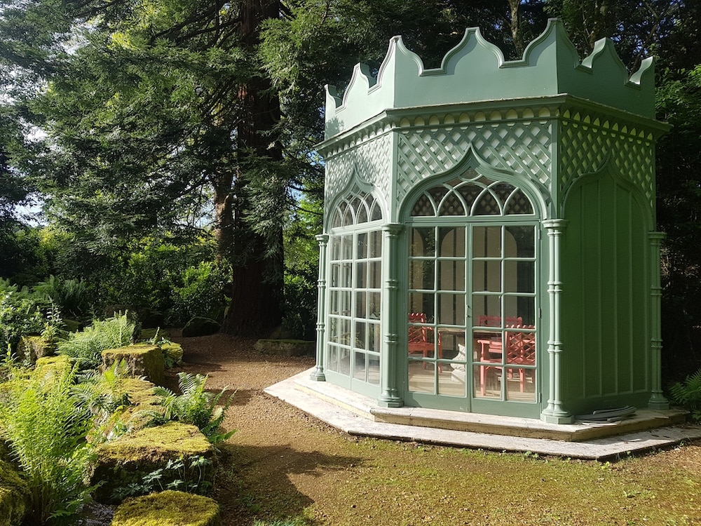 A green, decorative garden pavilion with large windows stands among trees and mossy rocks in bright sunlight.