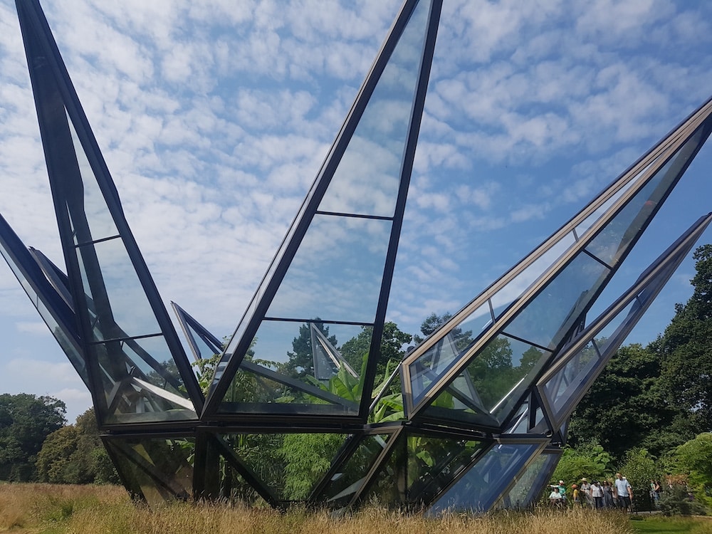 A large structure made of glass and metal with pointed panels stands on grass beneath a partly cloudy sky.