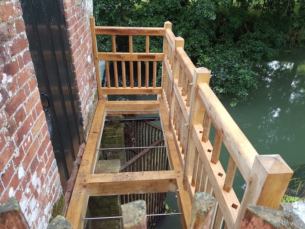 A wooden balcony with railings faces water beside a brick wall and a closed black door.