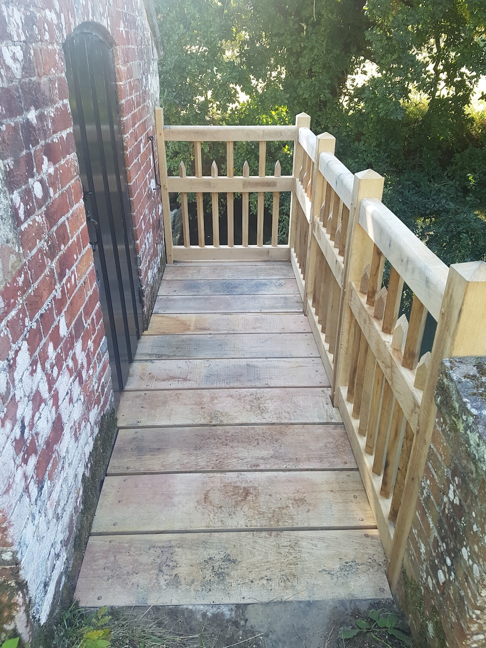 A wooden decking with a railing stands next to a brick wall, providing a view of green trees and grass.
