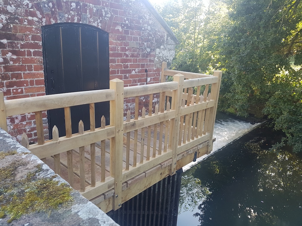 A wooden balcony with a picket railing looks out over a flowing stream next to a red brick building.