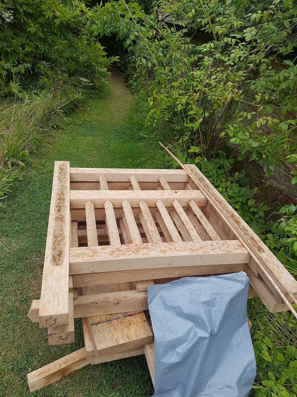 Wooden planks and beams are stacked on grass beside a garden path, partially covered by a grey sheet.