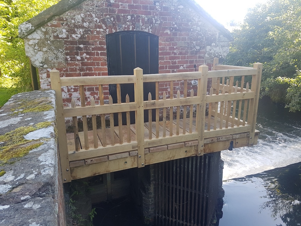 An old brick building with a dark door stands behind a wooden balcony that extends over the water.