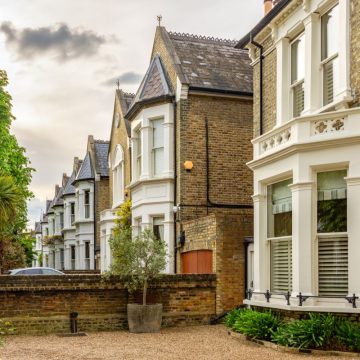 White terraced houses with black railings, window balconies, and potted plants line a sunny residential street.